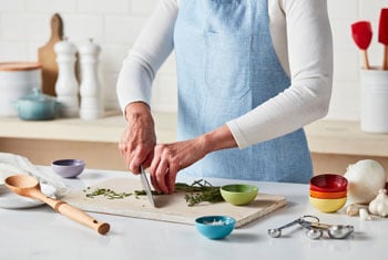 a woman chops herbs alongside prep bowls while wearing a blue kitchen apron