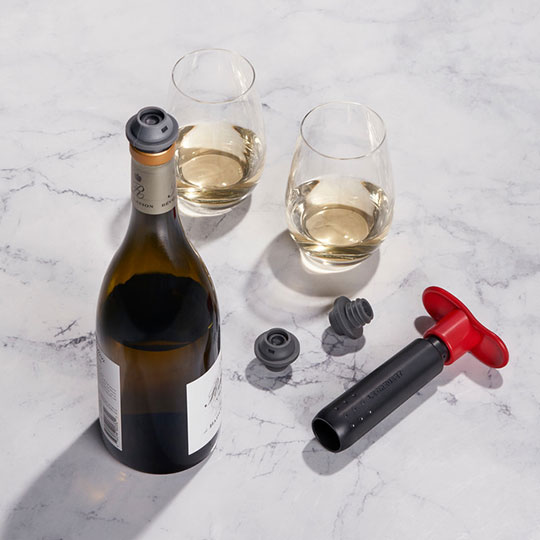 bottle of white wine alongside a black and red Le Creuset handheld wine pump and gray stoppers