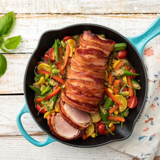pork loin wrapped in bacon atop veggies in a skillet